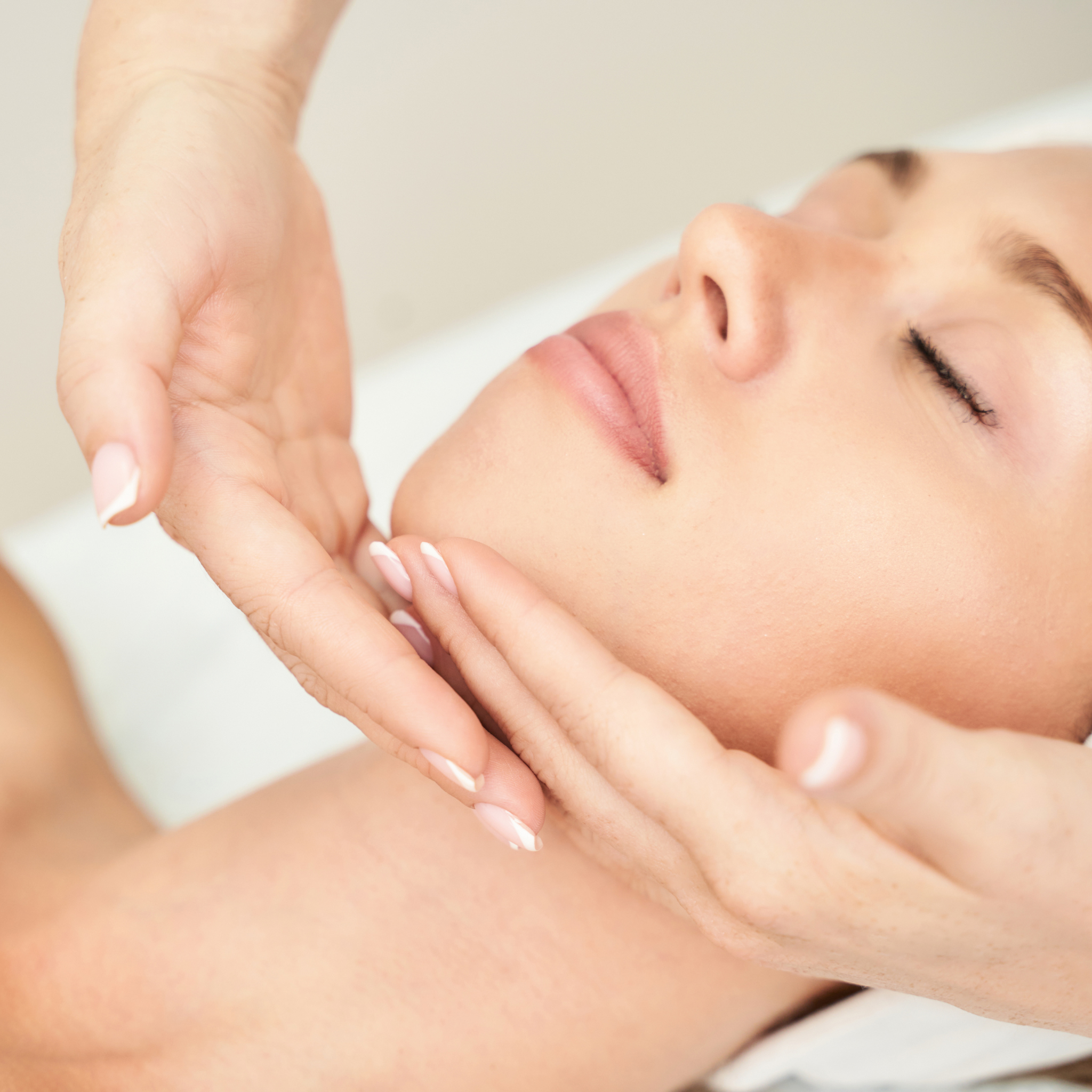 A close-up shot of a woman receiving a facial massage, with her face resting on a white surface and her hands gently cradling her jaw and cheek.