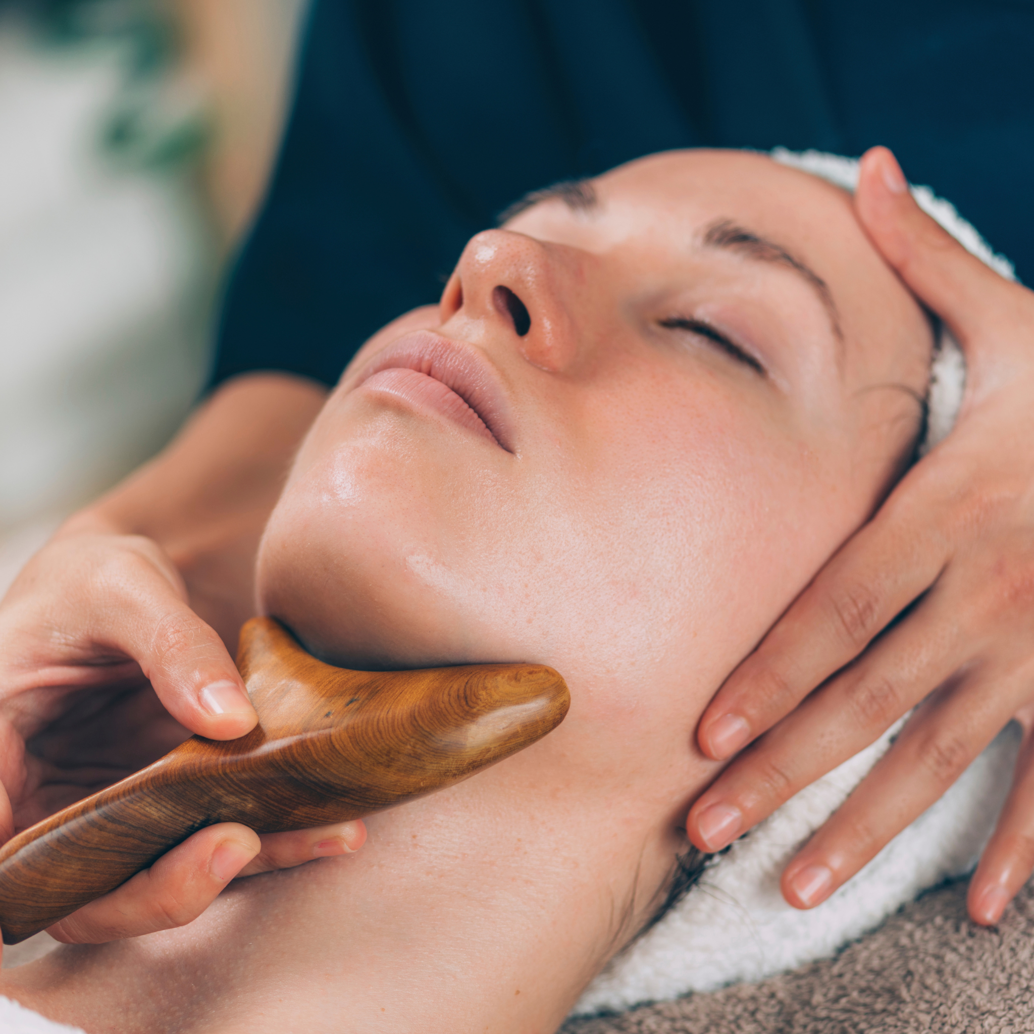 A close-up shot of a woman receiving a neck massage, lying with her eyes closed while a therapist uses a wooden gua sha tool on her neck. The therapist's hands and dark blue top are visible, along with a white towel wrapped around the woman's head.