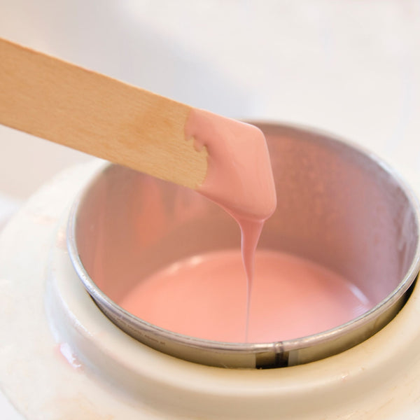 Pink wax being dripped from a wooden spatula into a white container.