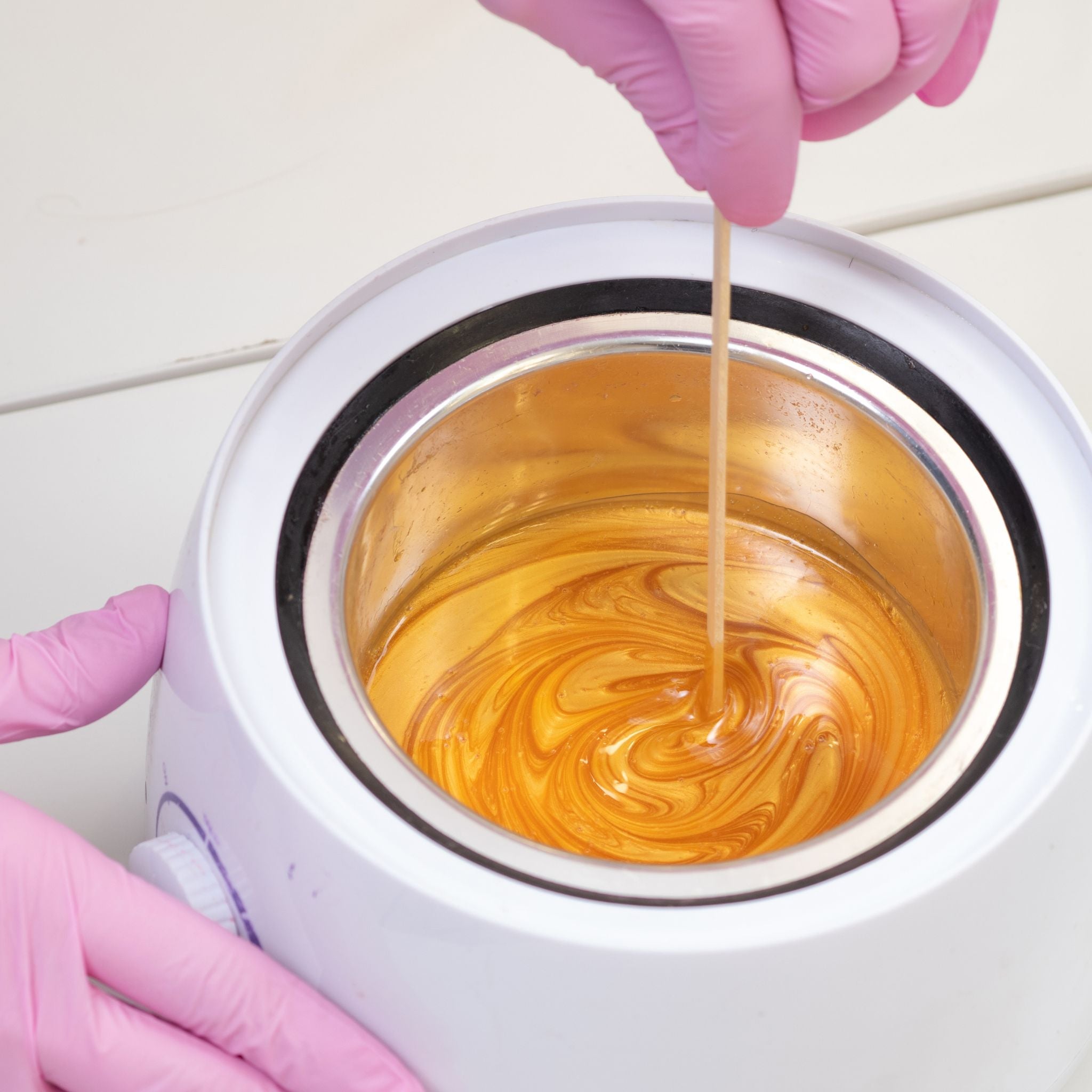 Person wearing pink gloves pouring wax into a white container with a swirling pattern.